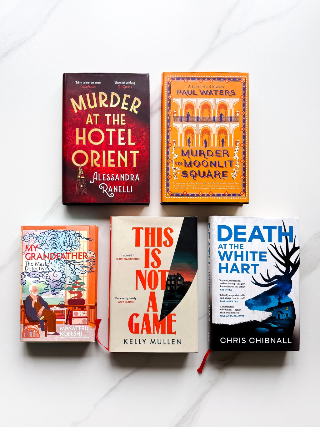 A flat lay photo of 5 cosy crime books: Murder at the Hotel Orient, Murder in Moonlit Square, My Grandfather the Master Detective, This Is Not a Game, Death at the White Hart